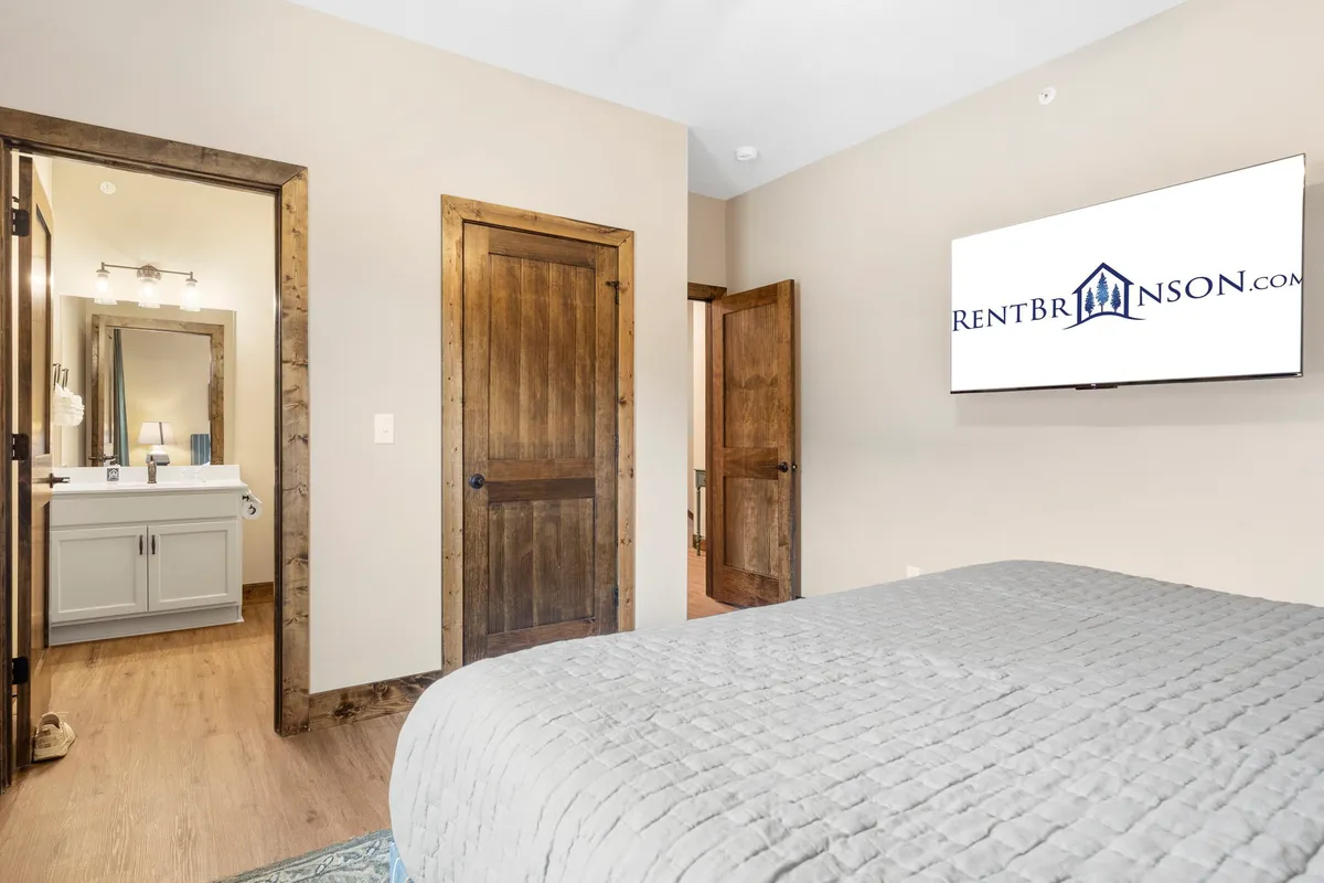 A cozy bedroom featuring a bed with textured bedding, softly lit by natural light filtering through the windows. Doorways lead to other rooms, and a wall-mounted television adds a modern touch, creating a peaceful and private space for relaxation.