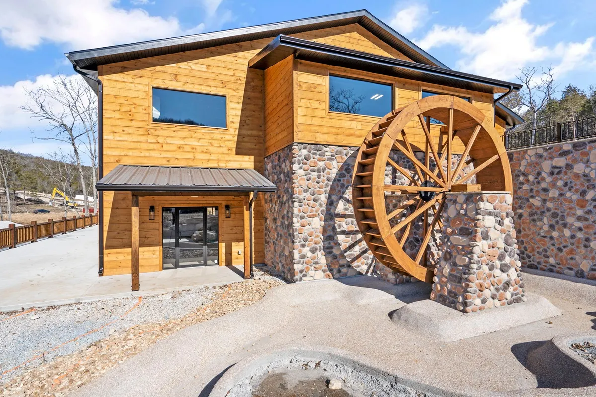A rustic water wheel set against a stone wall and wooden facade, surrounded by trees and inviting outdoor space. The scene suggests a historic, charming environment typical of the Ozarks and evokes a sense of local heritage and exploration.