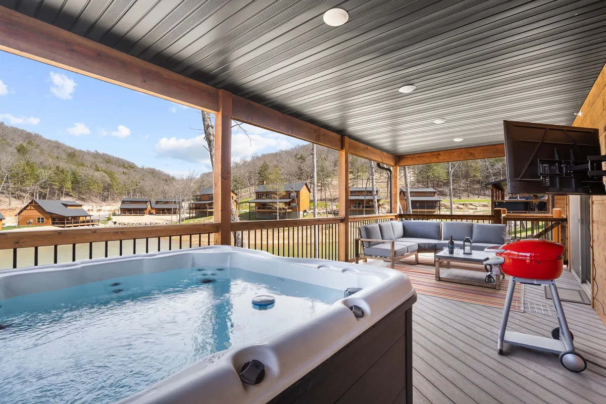 This exterior scene captures a wooden deck featuring a private hot tub, an outdoor seating area, a grill, and a television. The deck overlooks serene natural surroundings, possibly with water or sunset views in the distance. The setting is both inviting and intimate, suggesting the perfect spot for relaxation, stargazing, or socializing after a day outdoors.