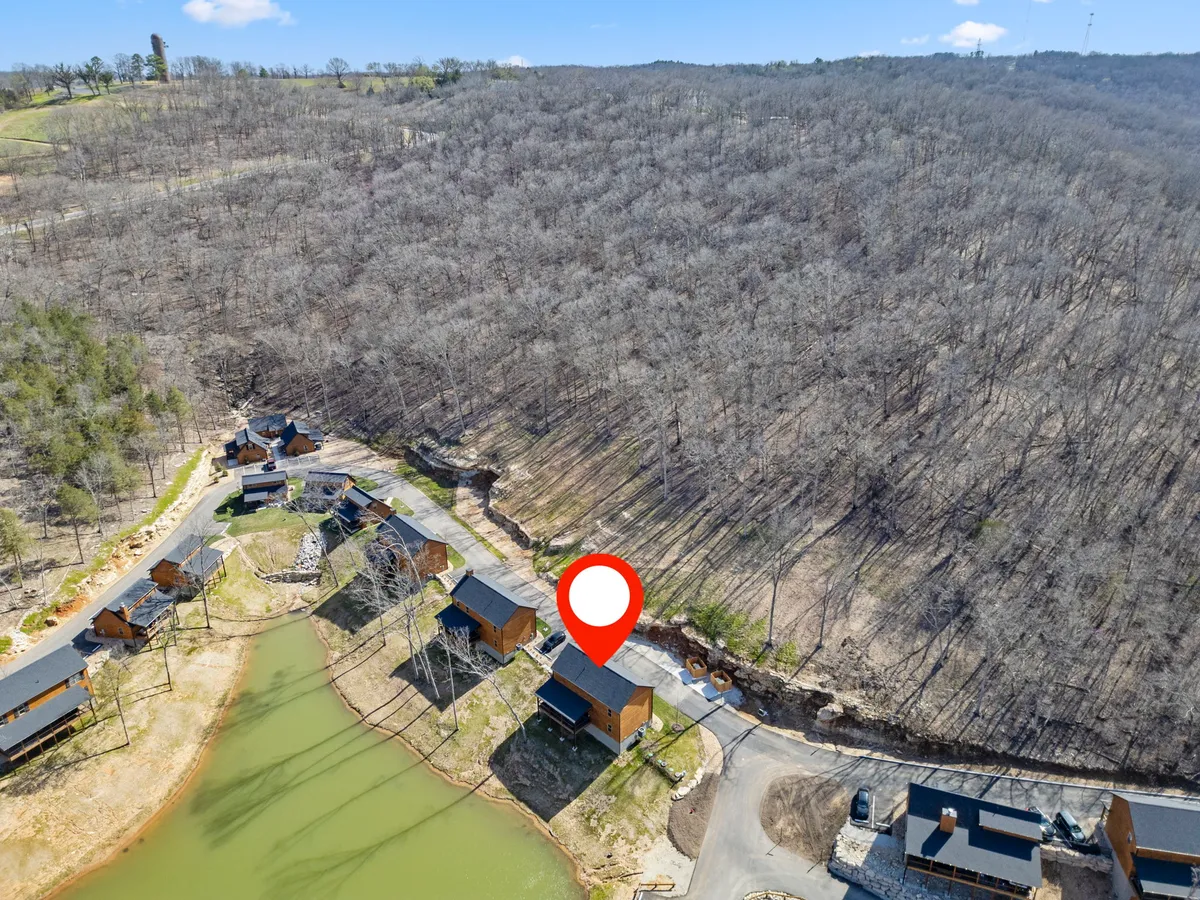 An exterior view featuring several cozy cabins nestled beside a tranquil pond, surrounded by forest and walking paths. The open space and the reflection of trees on the water evoke a peaceful, nature-oriented environment, perfect for outdoor adventures with pets.
