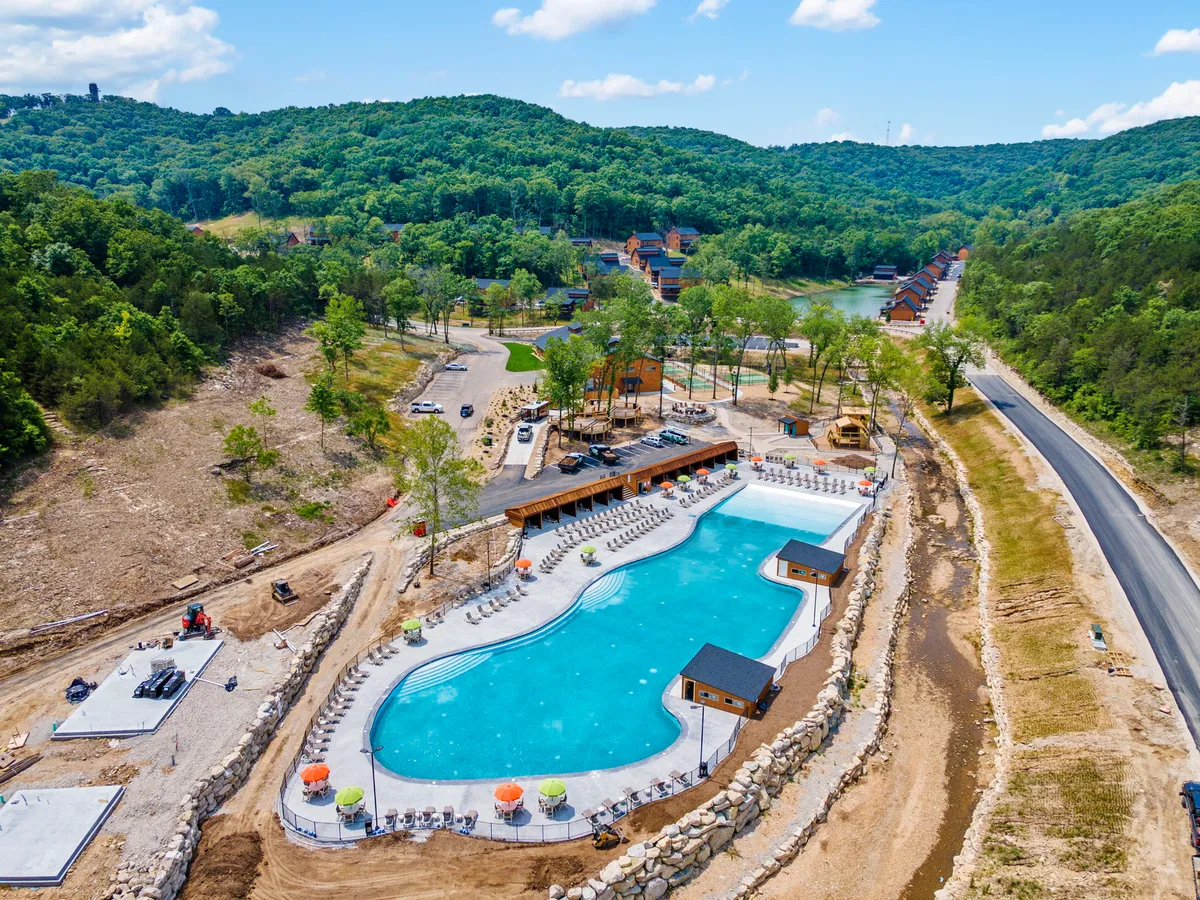 A vibrant outdoor area featuring a swimming pool surrounded by lounge chairs, trees, cabana structures, and a pathway leading towards cozy cabins. The pool area is inviting, with plenty of space for swimming, sunbathing, and gathering as a family.