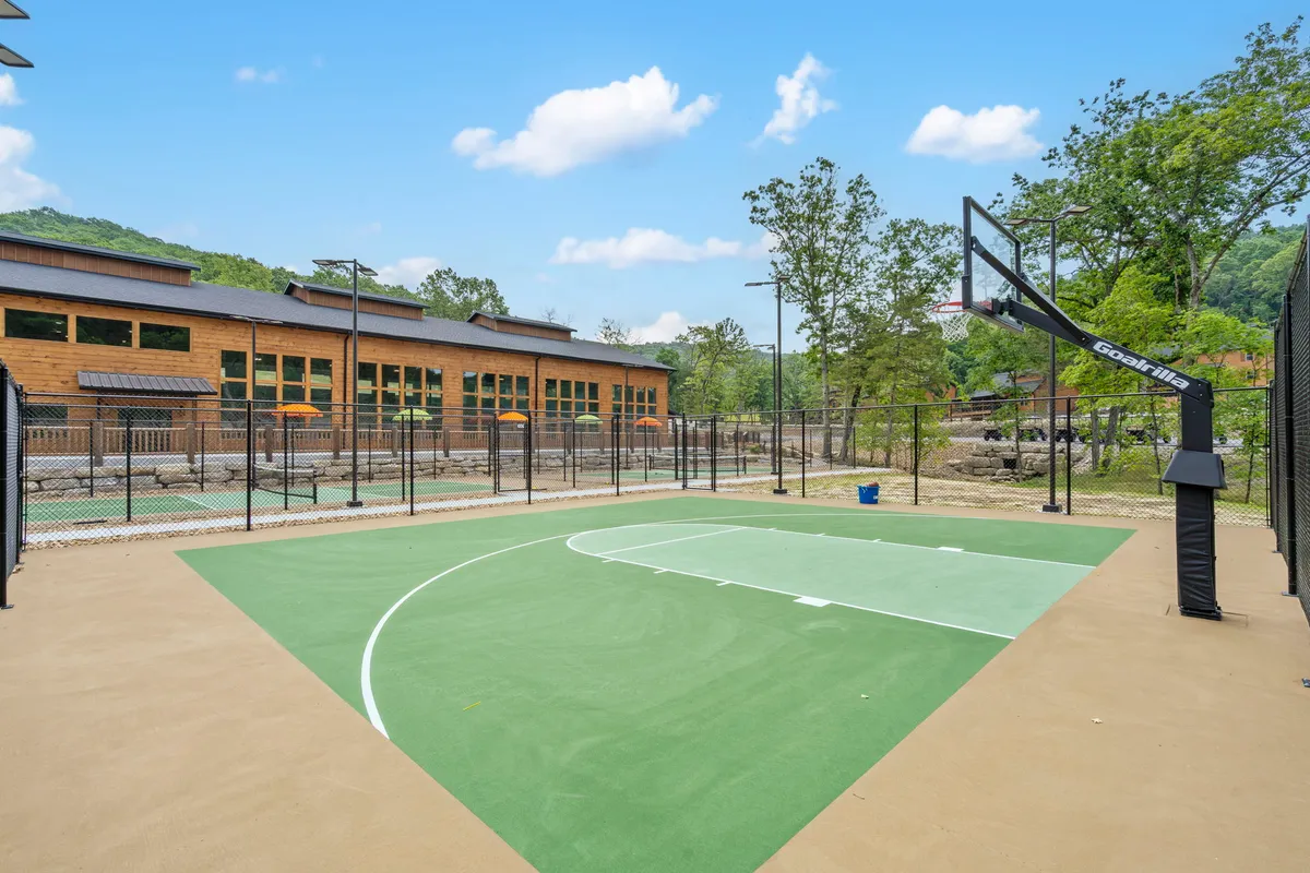An exterior view of a basketball court surrounded by trees and adjacent to a building. The well-maintained court invites families and groups to enjoy friendly matches and outdoor exercise while immersed in nature.