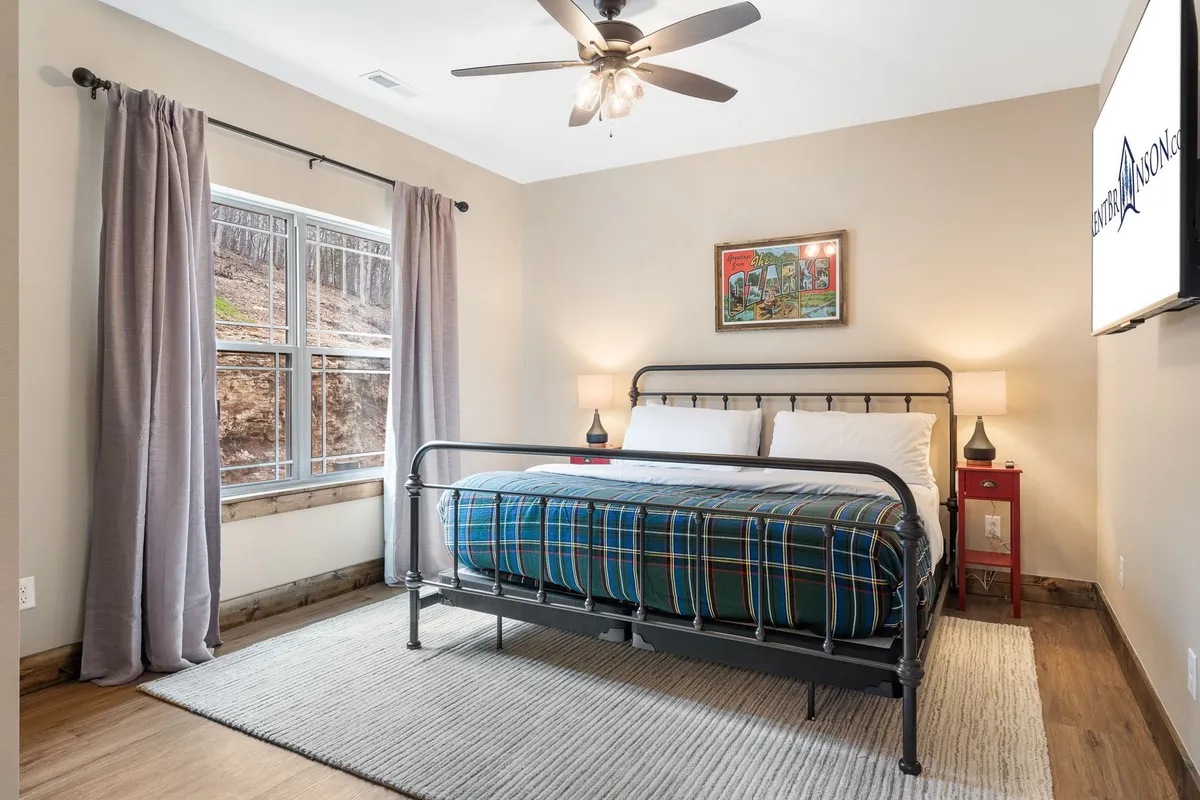 An inviting bedroom with a plaid blanket on the bed, a window allowing in gentle morning light, subtle artwork on the wall, and a nightstand with a lamp. The space feels warm and restful, perfect for recharging after a busy day.