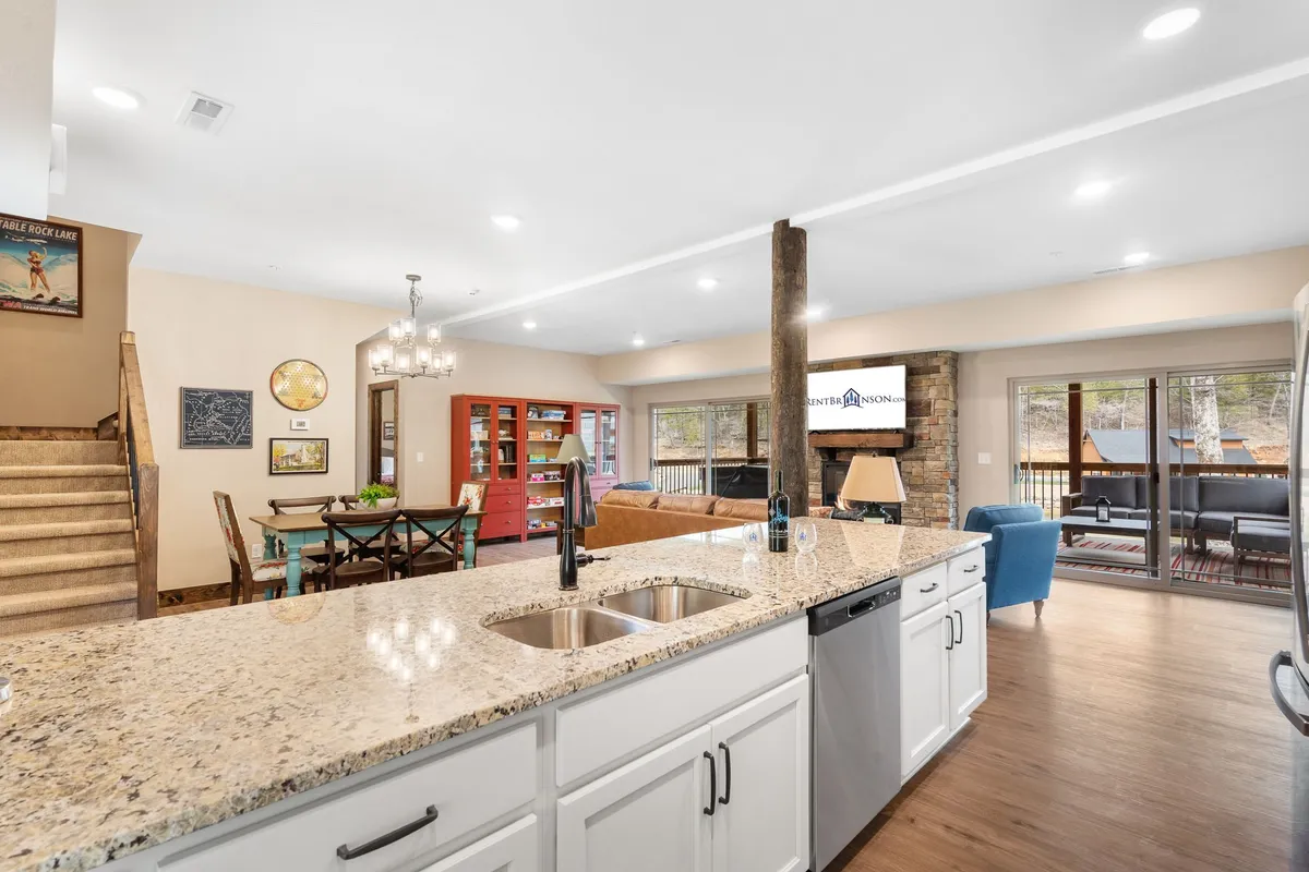 This image displays a welcoming open-concept interior featuring a granite countertop, kitchen island, wooden beams, dining area, and large windows that fill the space with light. The layout brings together the kitchen, dining, and lounging areas in one cohesive view.