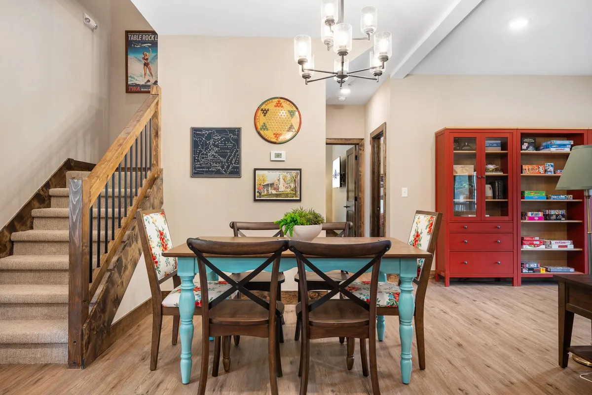 The image showcases a beautifully lit, open-concept dining area featuring a spacious wooden table perfect for group gatherings, a striking chandelier casting a warm glow, an eye-catching red cabinet, wall art that adds character, and a staircase hinting at a multi-level retreat. The inviting ambiance suggests a space designed for memorable shared meals, lively conversations, and cherished family celebrations.
