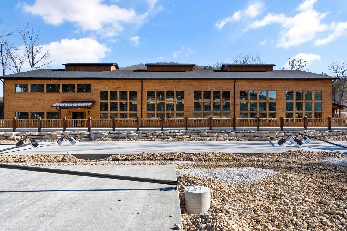 A welcoming exterior shot featuring a wooden building with large windows and a stone wall, surrounded by greenery. The open patio space is ideal for gathering, relaxing, or enjoying sunset views, and visually ties the home to its beautiful surroundings.