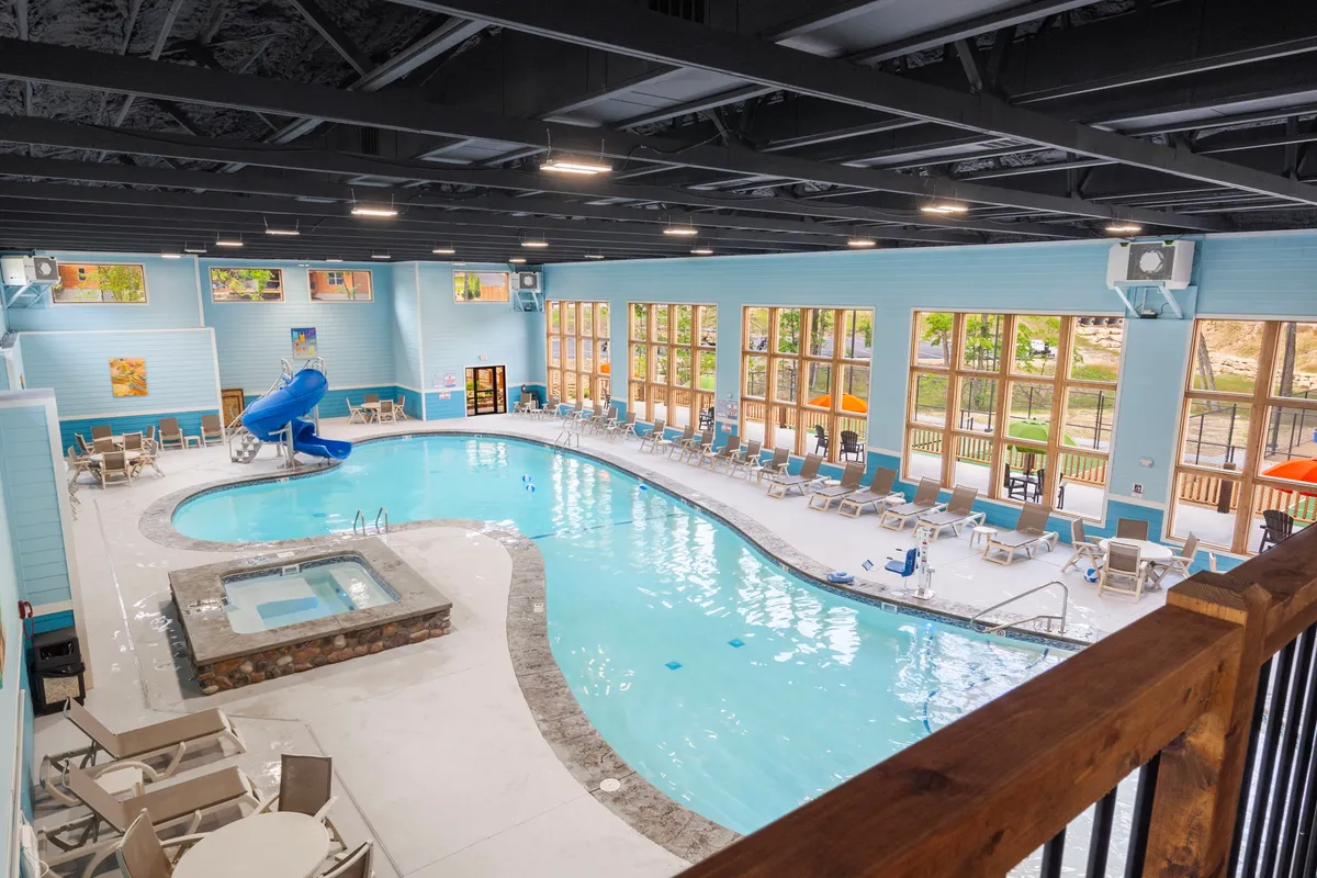This interior image features a vibrant indoor swimming pool area with a water slide, hot tub, lounge chairs, and large windows letting in natural light. The mood is joyful and relaxing, with plenty of space for swimming, lounging, and family gatherings.